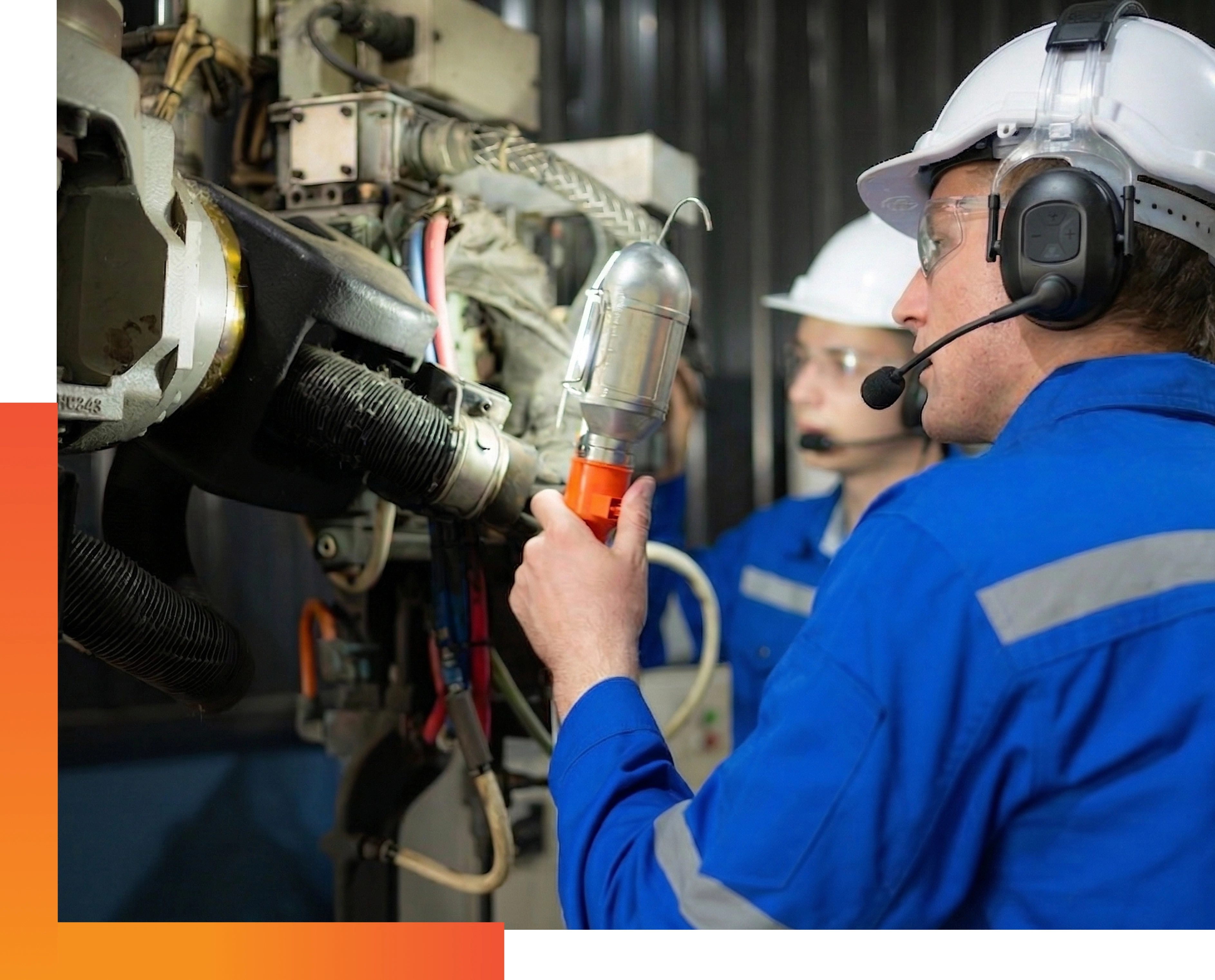 Two workers in blue uniforms and white helmets examine machiner holding a tool, while both wear headsets, showing focus and teamwork.